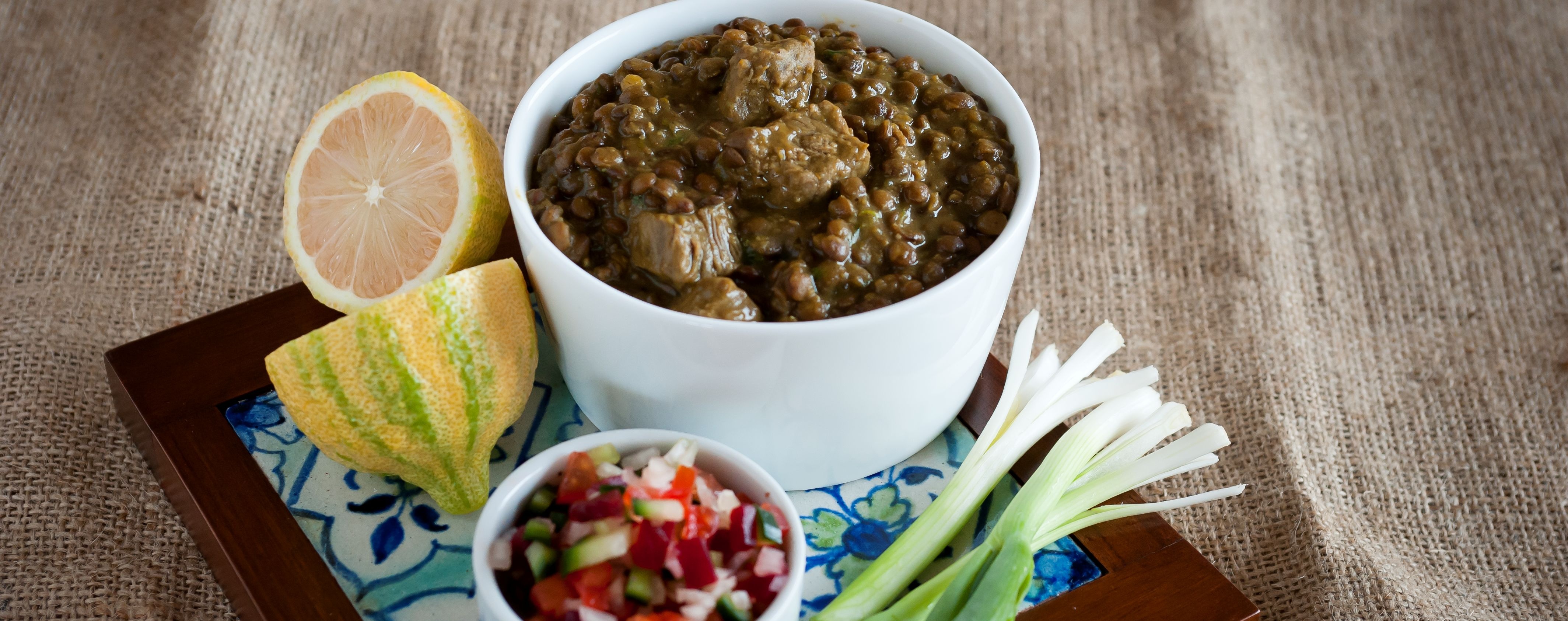 Ma gos, Parsi meat stew in a white china bowl, on a ceramic tile with a fresh chutney and halved citrus.