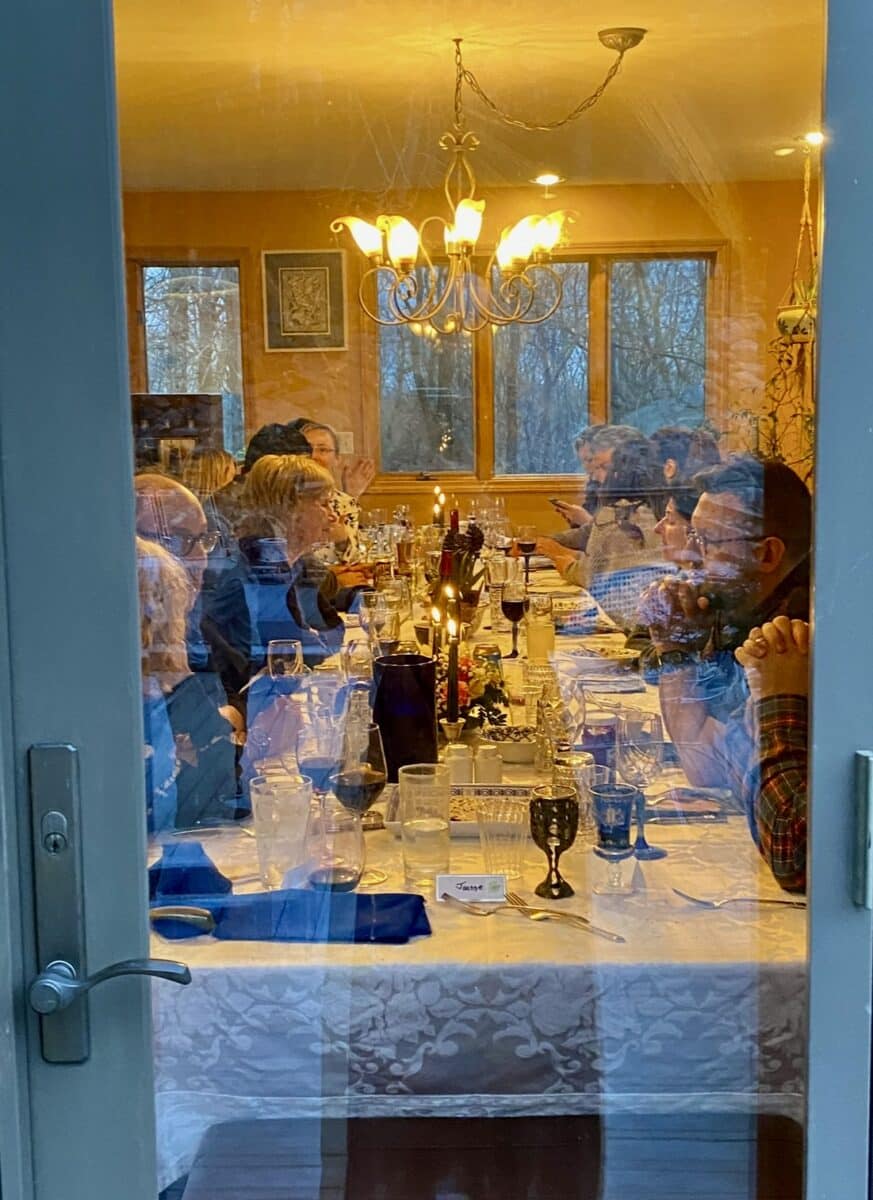 A Jewish family gathered around a Passover dinner table, as seen through a glass door from outside the home.