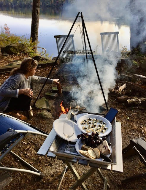 Woman tending a campfire by a lake, with a camp table in the foreground holding wine glasses, grapes and cheese.