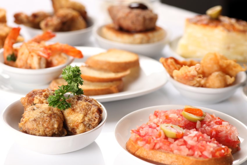 Array of assorted appetizers on white plates, including bread, meat, and fresh sides.