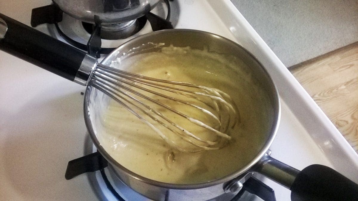 Whisk stirring a creamy béchamel sauce in a saucepan on a stovetop.