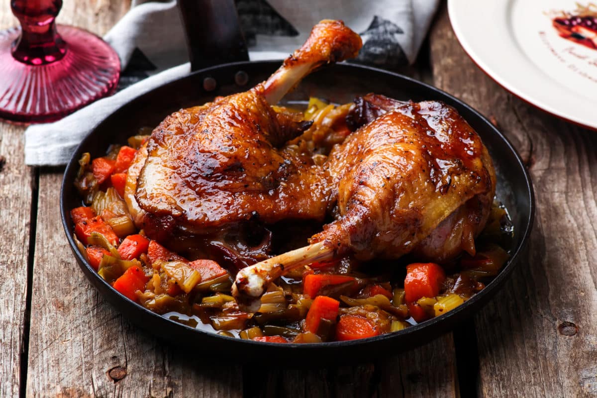 Braised duck legs and vegetables served in a skillet on a rustic wooden table.