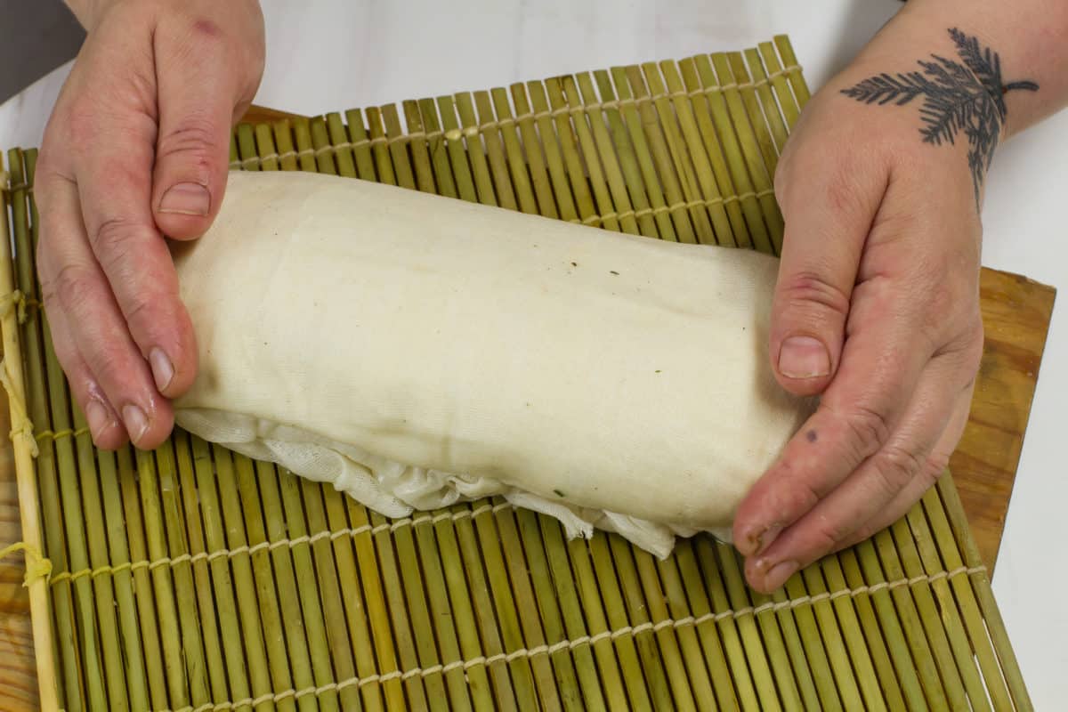 Close-up of two hands carefully shaping a cheese log on a bamboo mat.