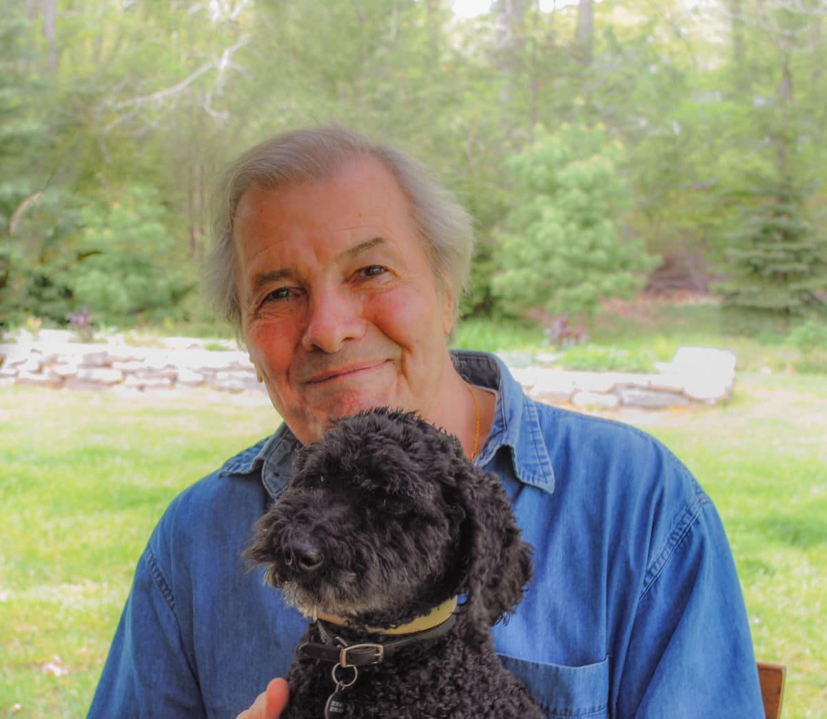Renowned Chef Jacques Pepin smiling with his black dog, Coco, in a natural setting.