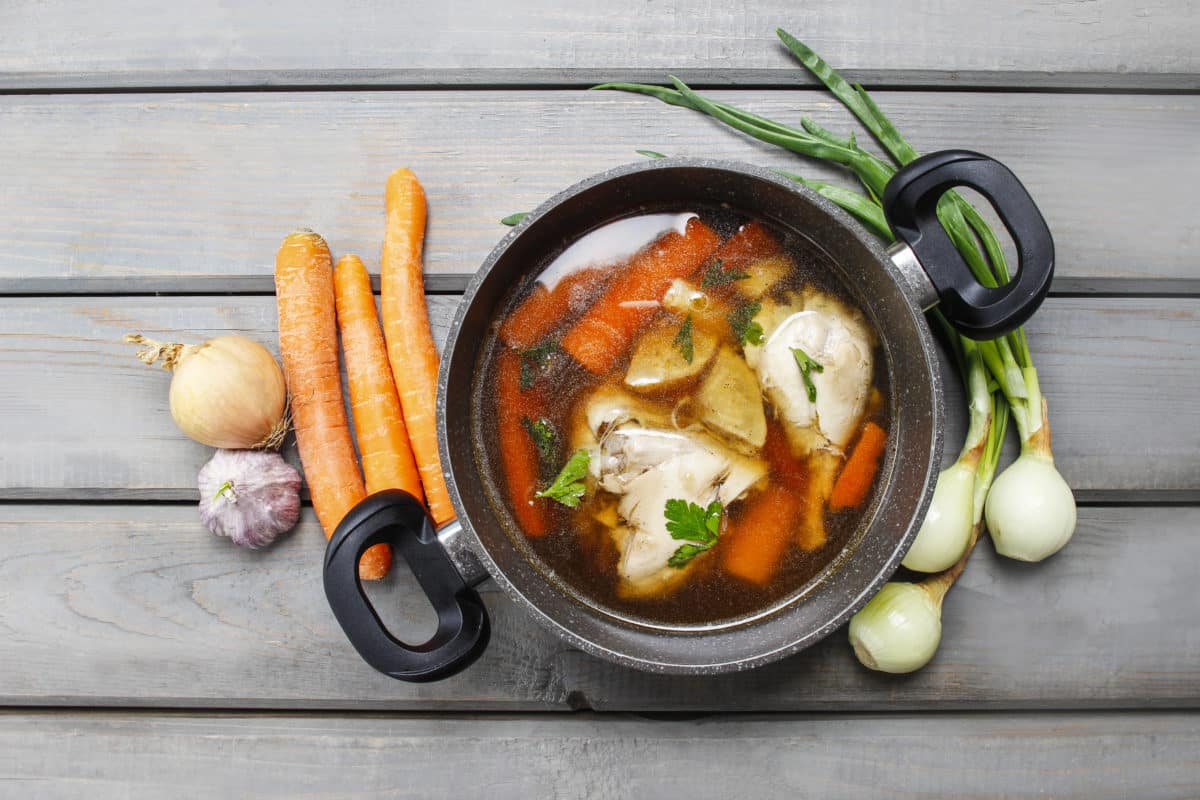 Pot of chicken soup with fresh vegetables on a wooden table.