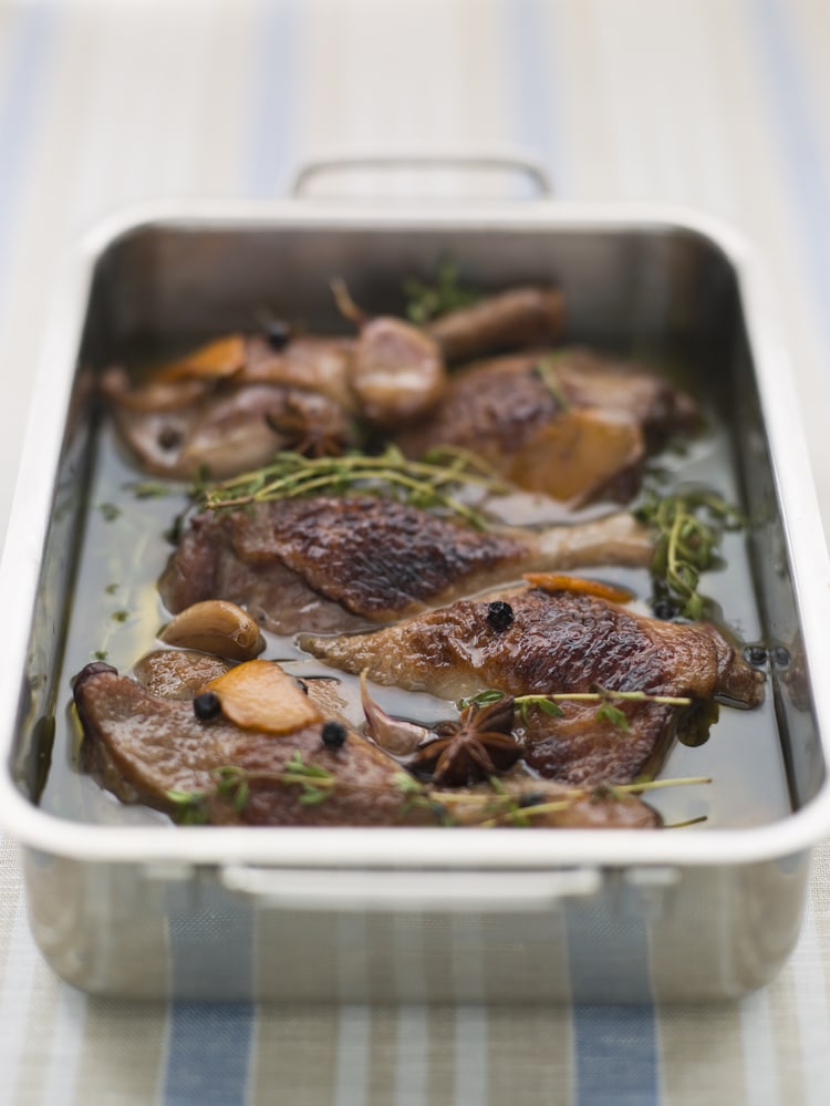 Cooked confit duck legs with herbs in a metal tray.