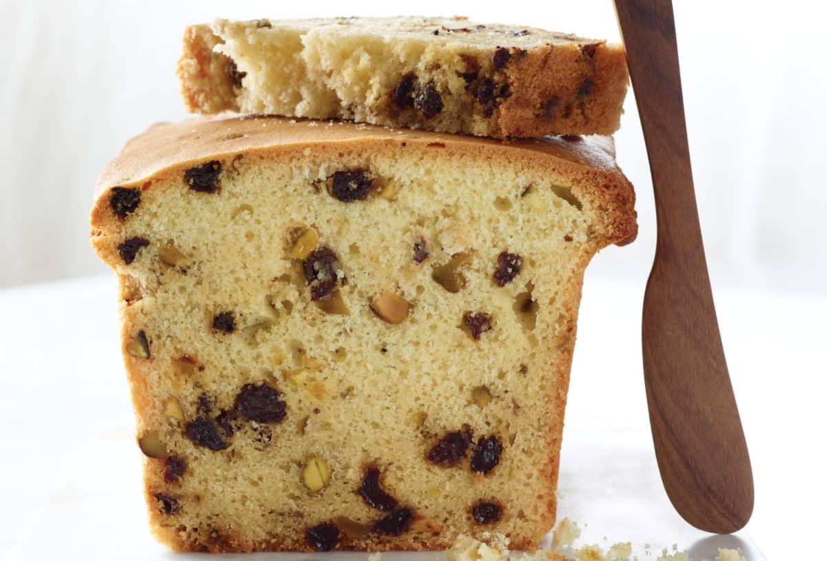 A sliced corn flour tea cake with currants, pistachios, and a wooden knife on the side.
