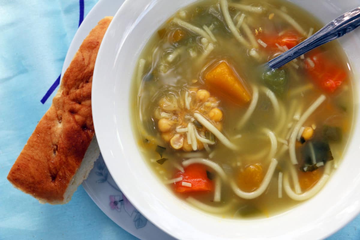 A close-up shot of a bowl of soup with fresh vegetables and noodles, accompanied by a slice of bread.