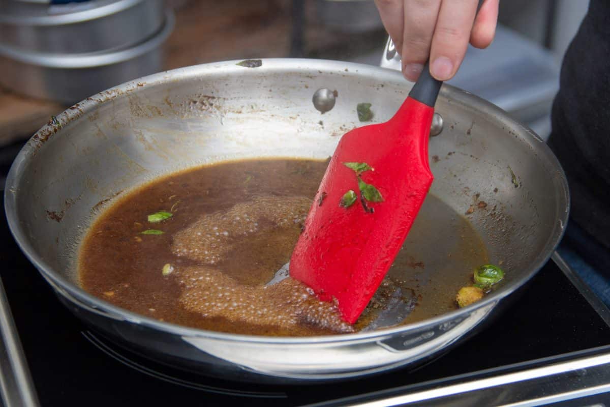 Hand using a red spatula to deglaze a stainless steel pan with brown sauce.