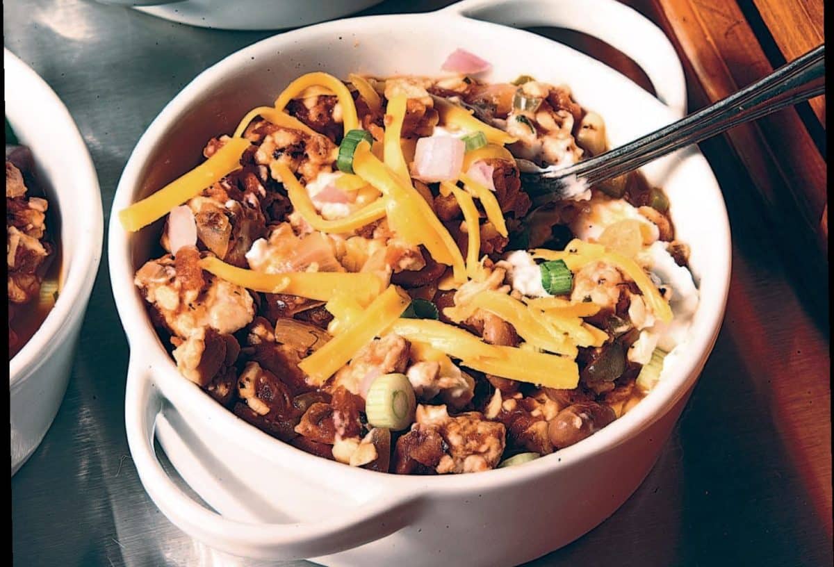White crock containing chili topped with cheese, onions, and other ingredients.