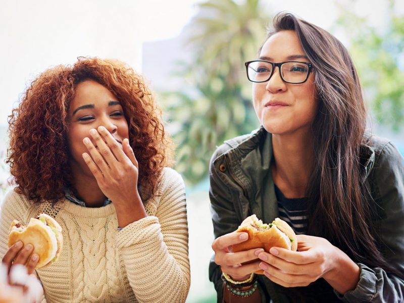 Two young women joyfully eating sandwiches outdoors.