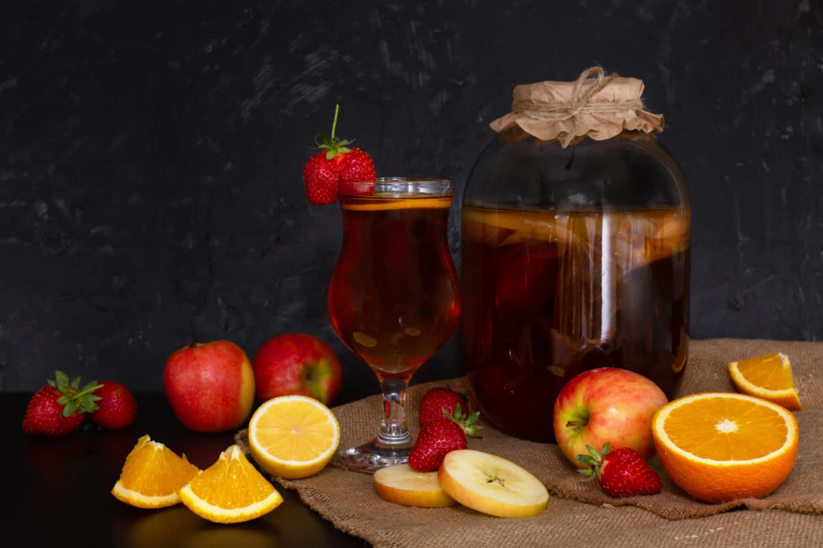 Glass of kombucha infused with various fruits alongside a brewing jar.