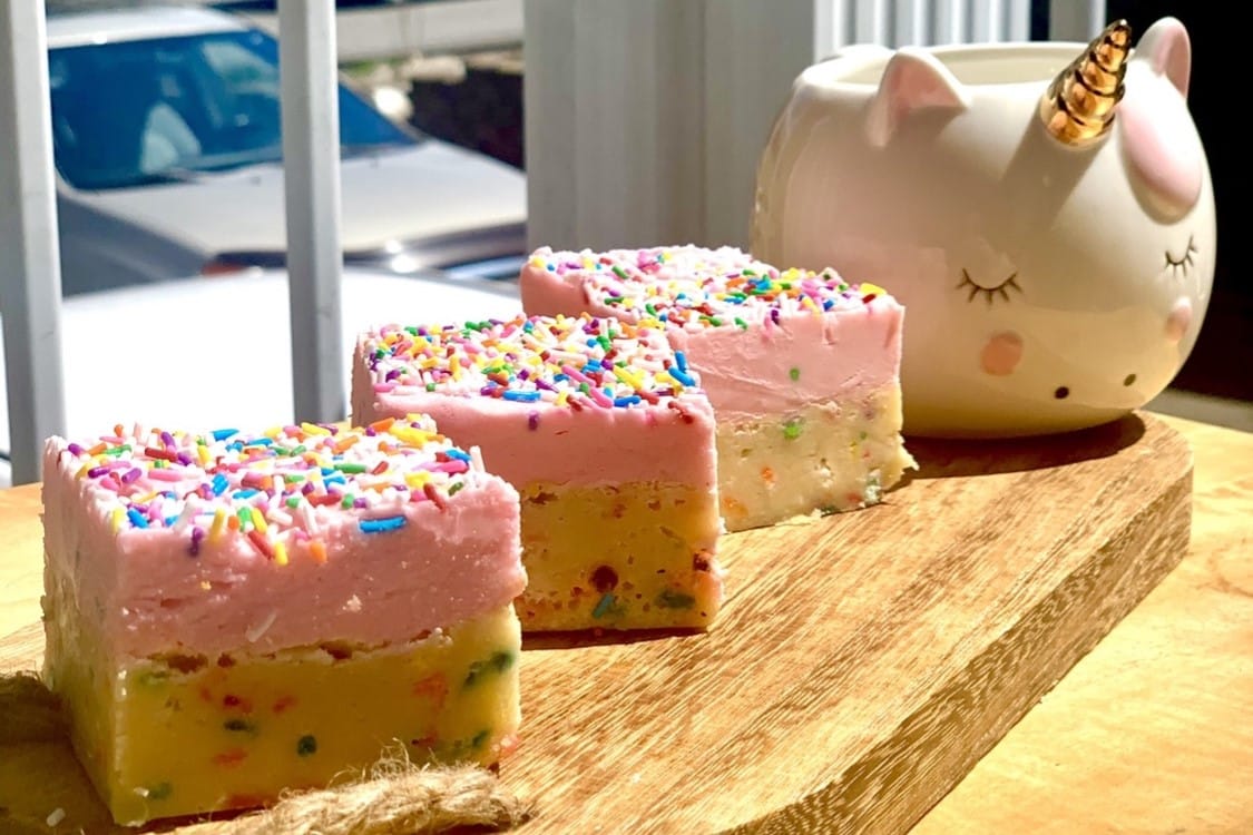 Close-up of funfetti blondies topped with pink buttercream and colorful sprinkles, next to a unicorn-themed container.