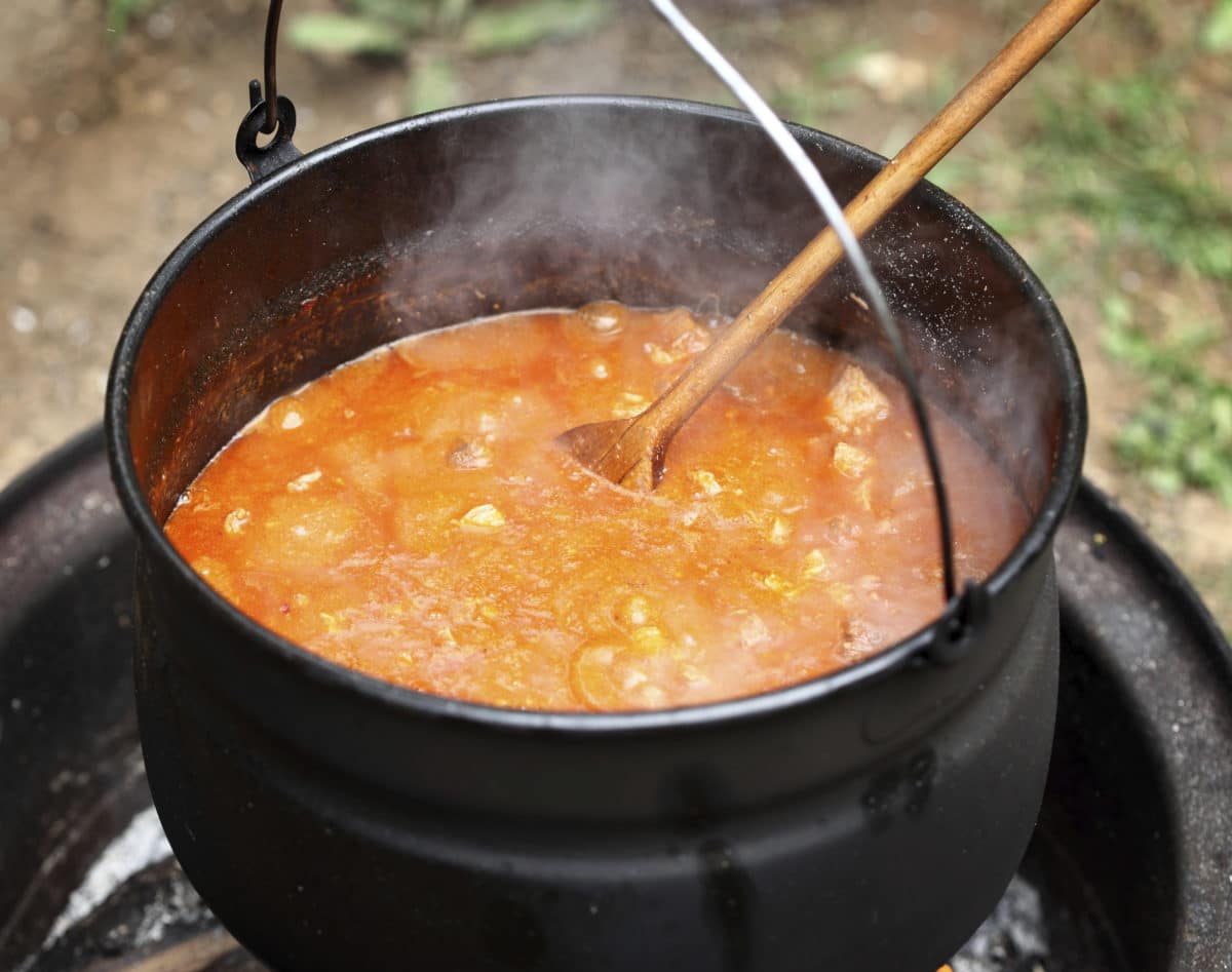 A pot of hobo stew cooking over an open flame with a wooden ladle stirring inside.