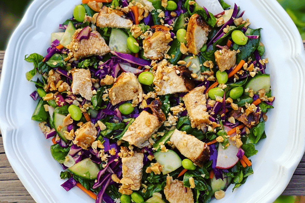 Fresh salad with hoisin glazed chicken, edamame, and assorted colorful veggies on a white plate.