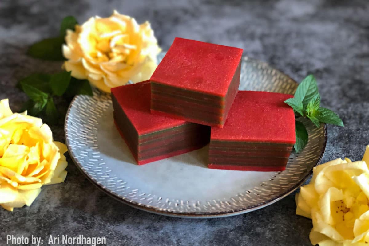 Stack of red Kuih Lapis, a layered coconut cake, on a silver plate with flowers.