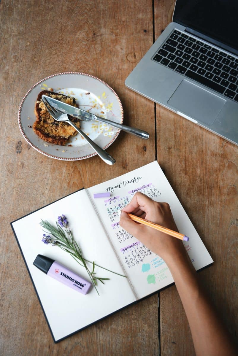 Laptop beside a plate of food on a wooden table with a person writing in a notebook.
