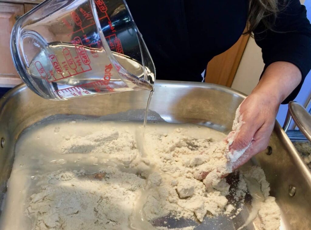 Close-up of a woman's hands adding water to flour to make pupusa dough in a silver mixing bowl.