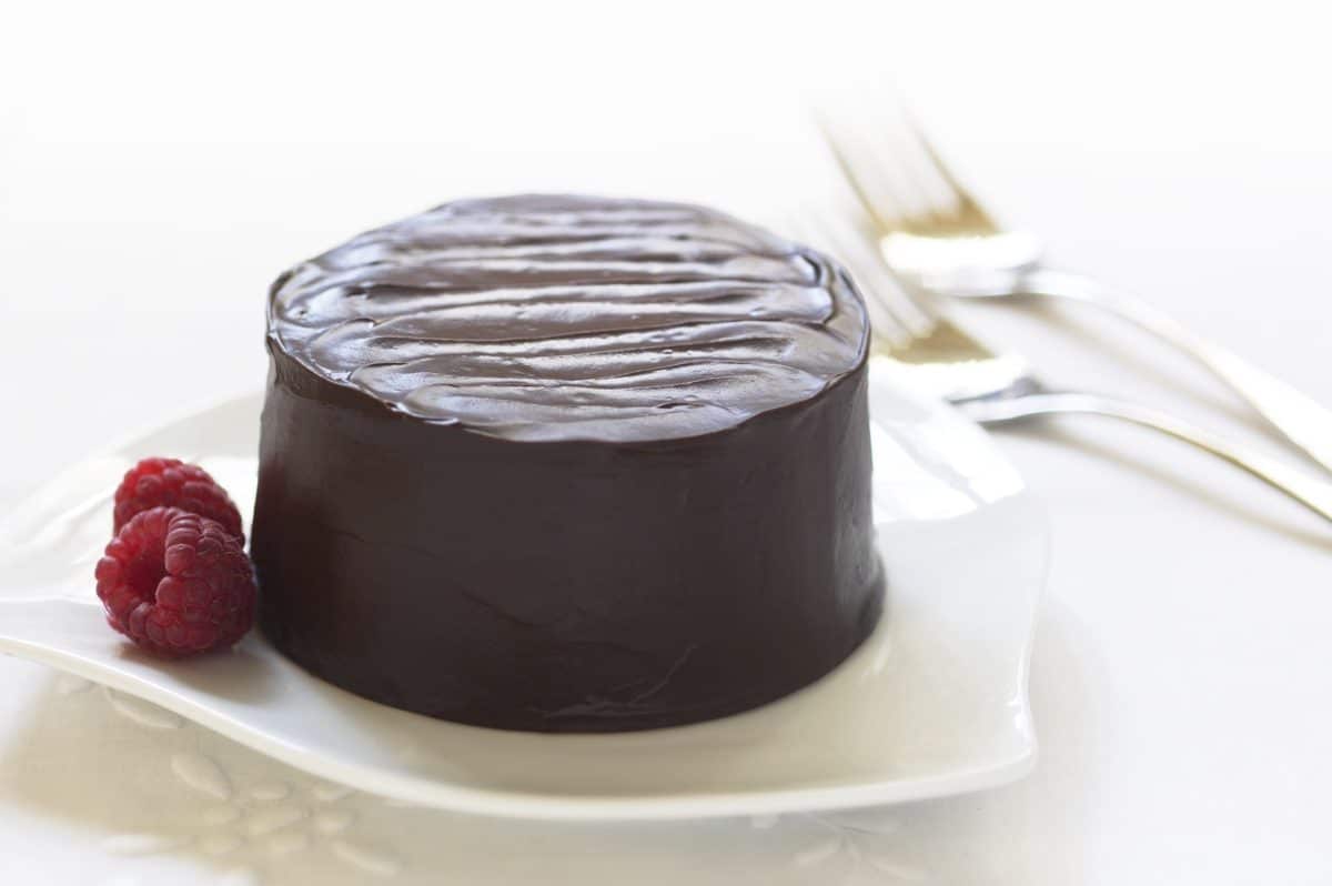 A glossy chocolate cake adorned with fresh raspberries on a white plate.