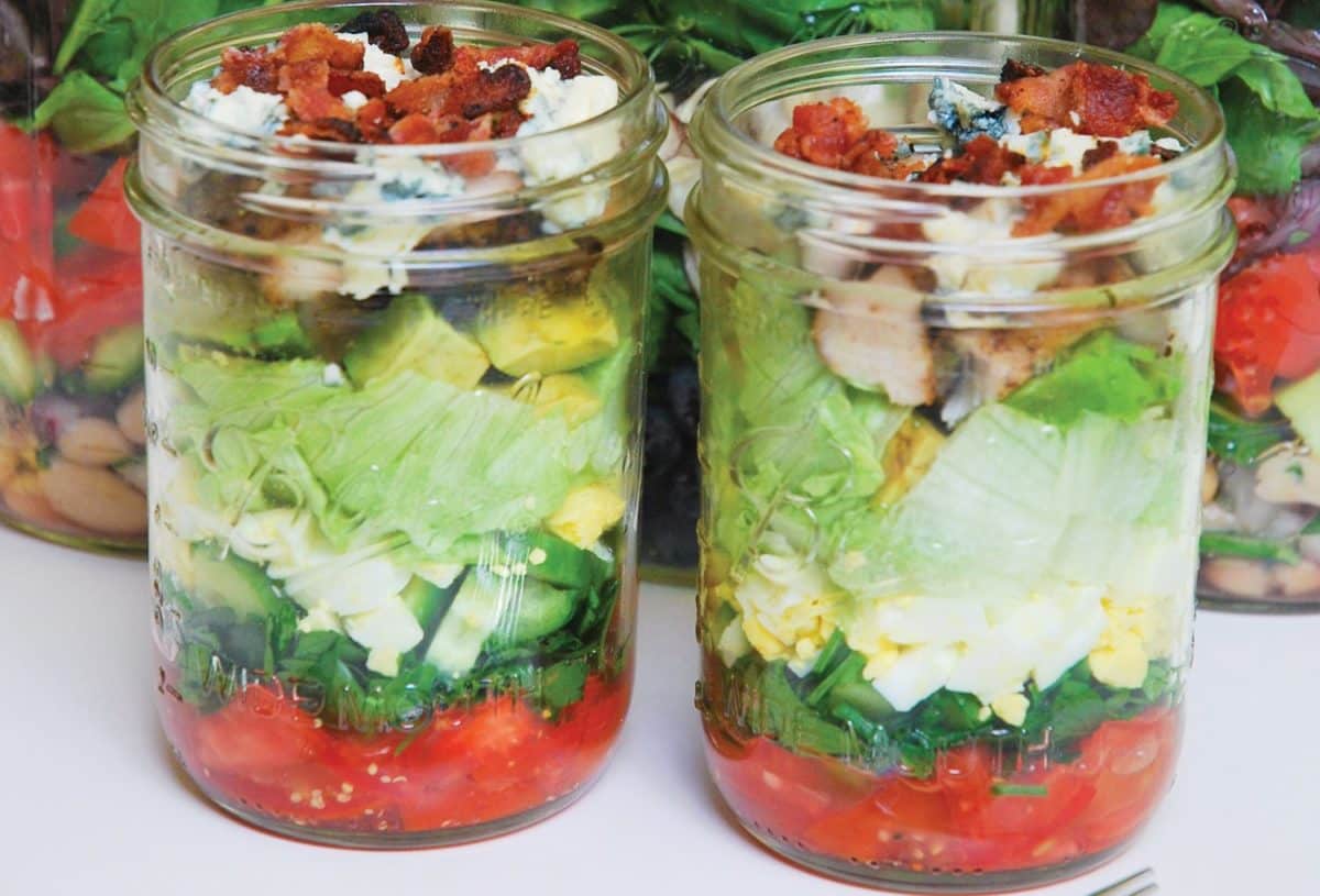 A layered Cobb Salad in two mason jars with ingredients like bacon, eggs, and vegetables.