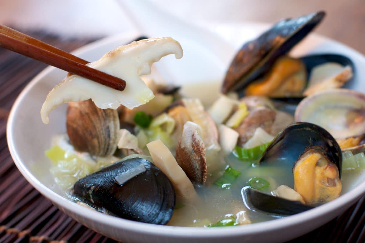 Steaming bowl filled with a clear broth, mushrooms, and various shellfish, with chopsticks lifting a piece of mushroom.
