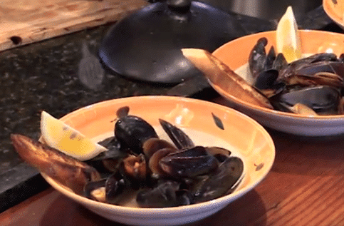 Two serving bowls of mussels steamed in ale accompanied by garlic bread and a lemon wedge.