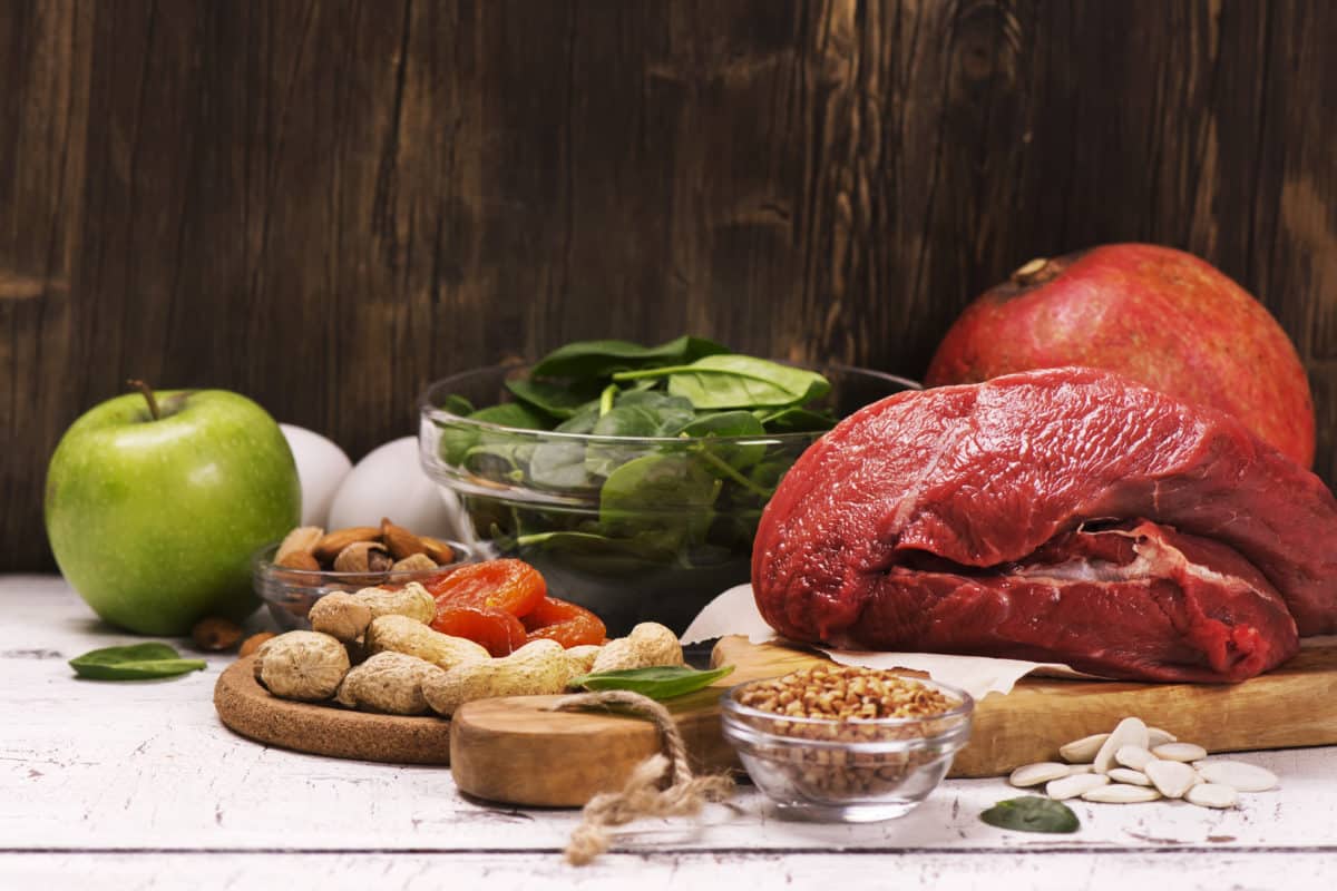 A variety of iron-rich foods including apple, spinach, red meat, pomegranate, seeds, and nuts.