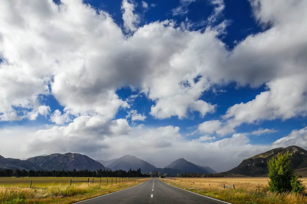A straight, empty road leading towards distant mountains under a deep blue sky scattered with large white clouds.