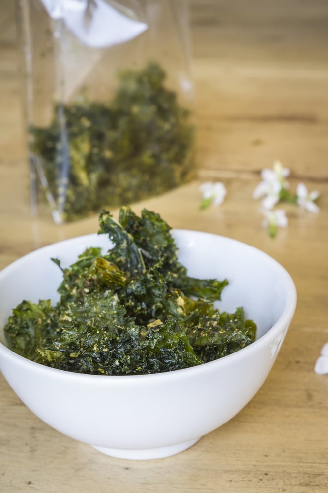 Kale chips seasoned with parmesan in a white bowl on a wooden table.