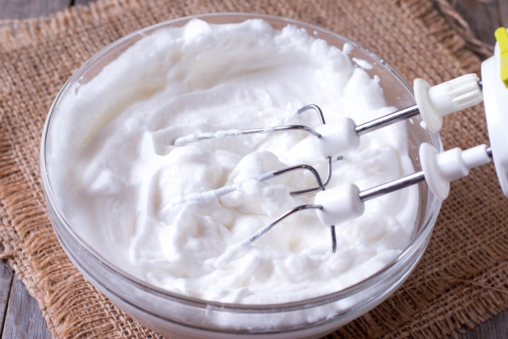 Whisk dipping into a bowl of fluffy whipped egg whites on a rustic wooden table.