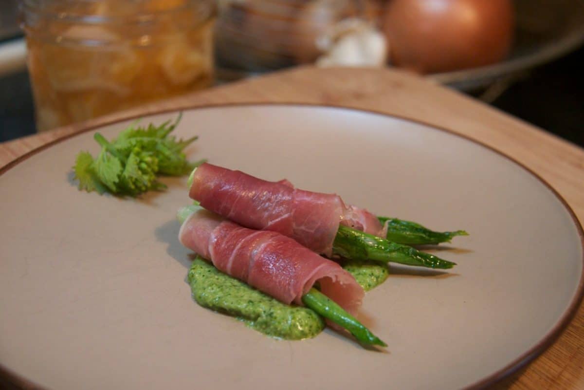 Hosta shoots wrapped in thinly sliced prosciutto, placed on a plate with garlic mustard pesto.