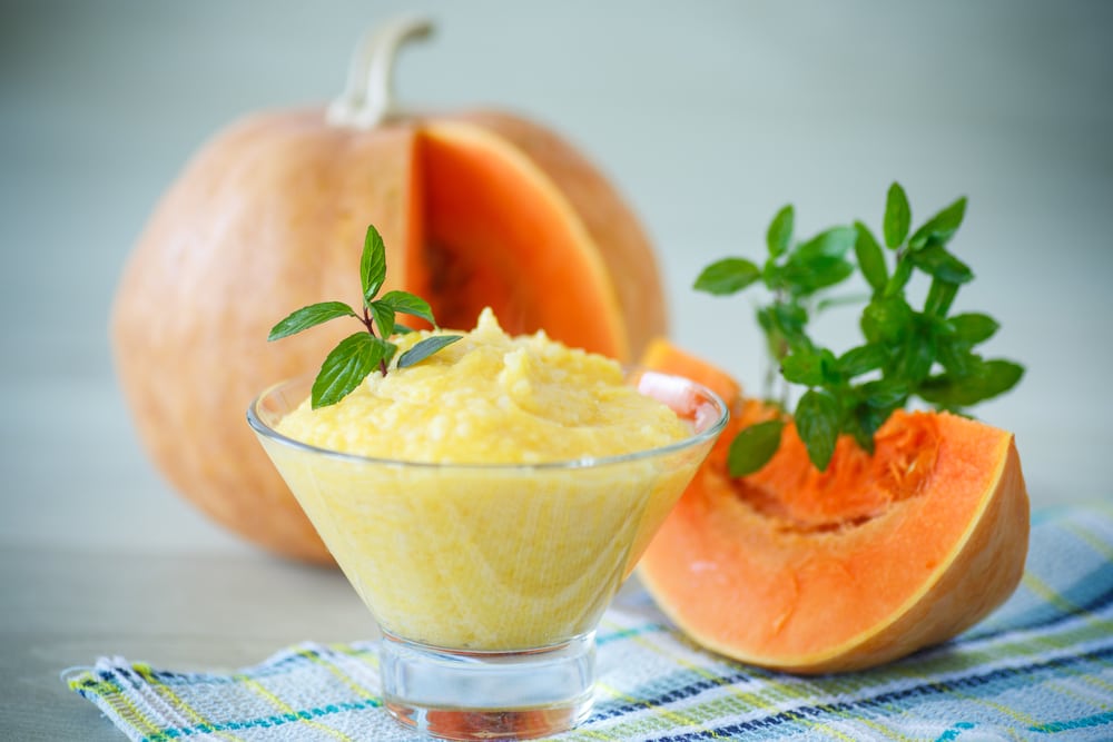 A glass bowl of pumpkin porridge garnished with mint against a backdrop of pumpkin