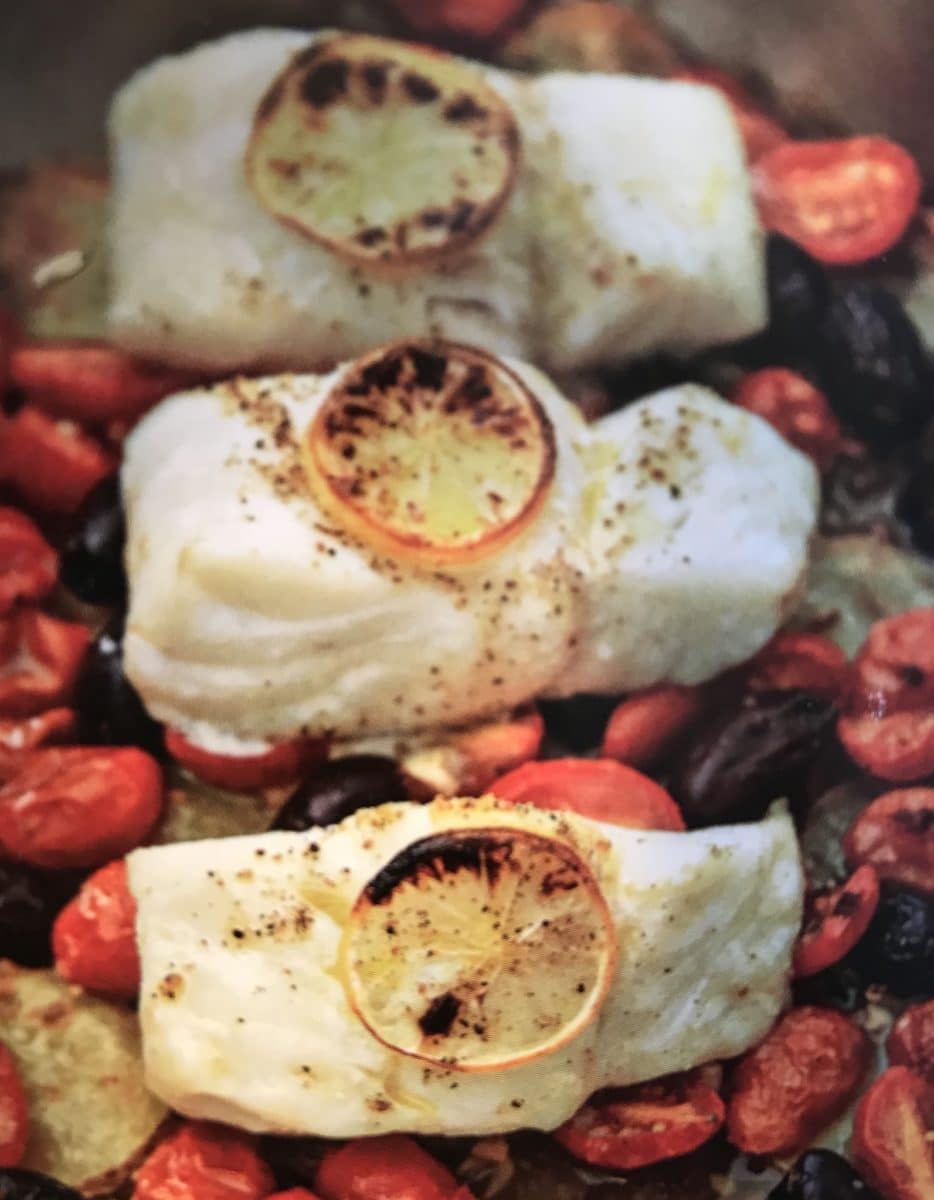 Roasted Cod served on a bed of roasted tomatoes, potatoes, and olives with slices of charred lemon on top.