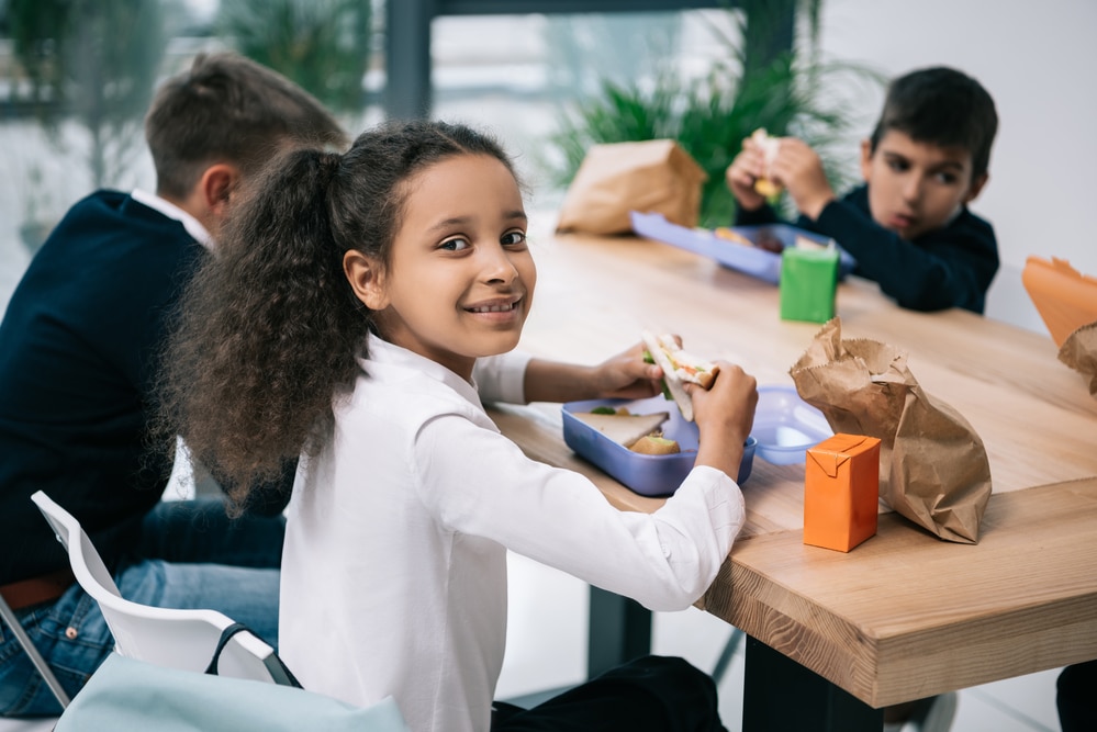 Kids sitting around a wooden school lunch table, eating lunches from home.