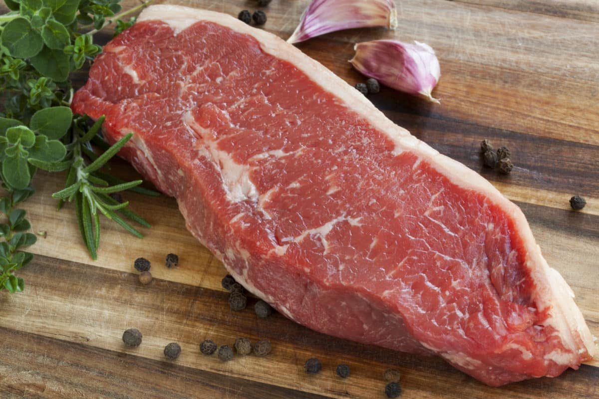 Raw sirloin steak on a wooden plank surrounded by garlic, peppercorns, and herbs