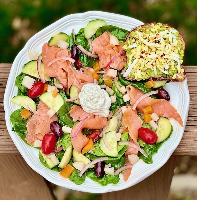A vibrant salad with smoked salmon, veggies, and an avocado toast on a white plate.