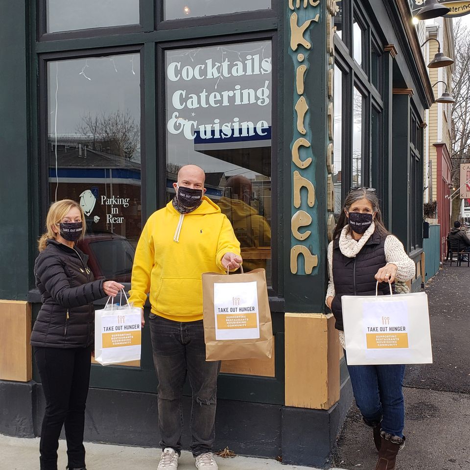 Three individuals in masks holding 'Take Out Hunger' bags outside a local eatery.
