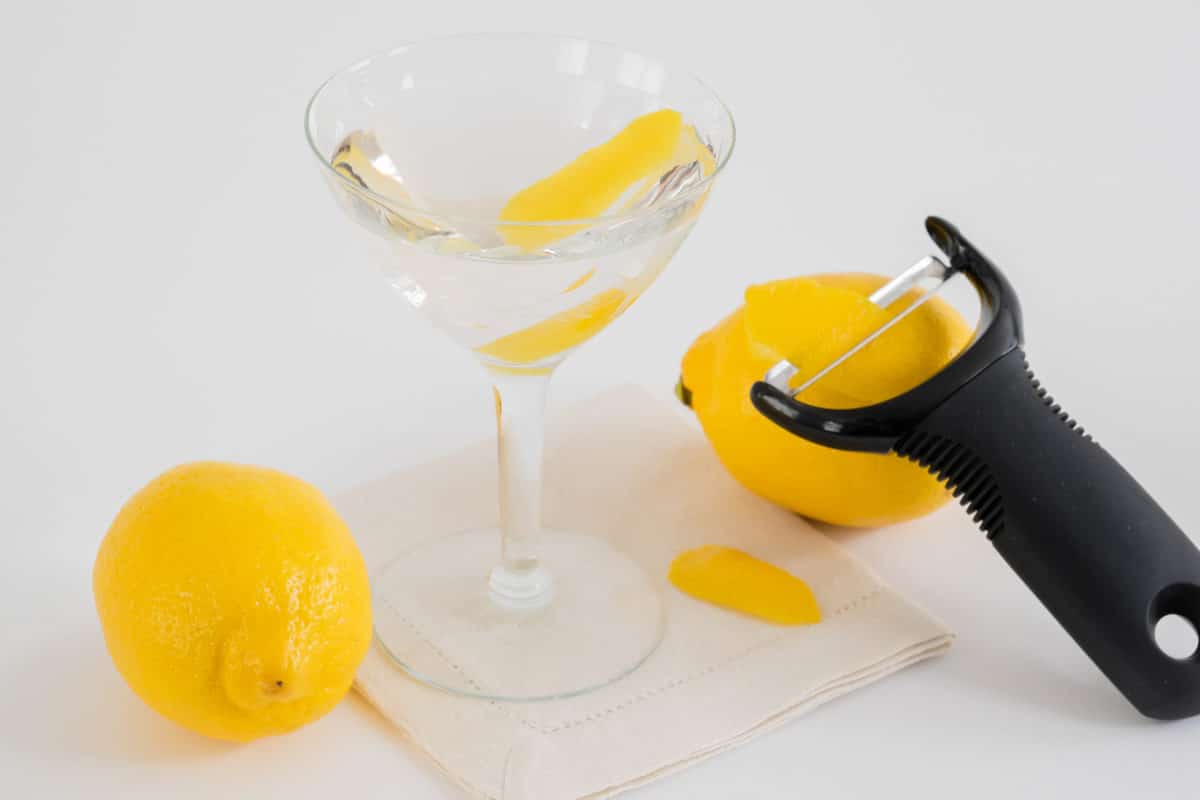 Martini glass with clear liquid and lemon peels, adjacent to a tool peeling a lemon