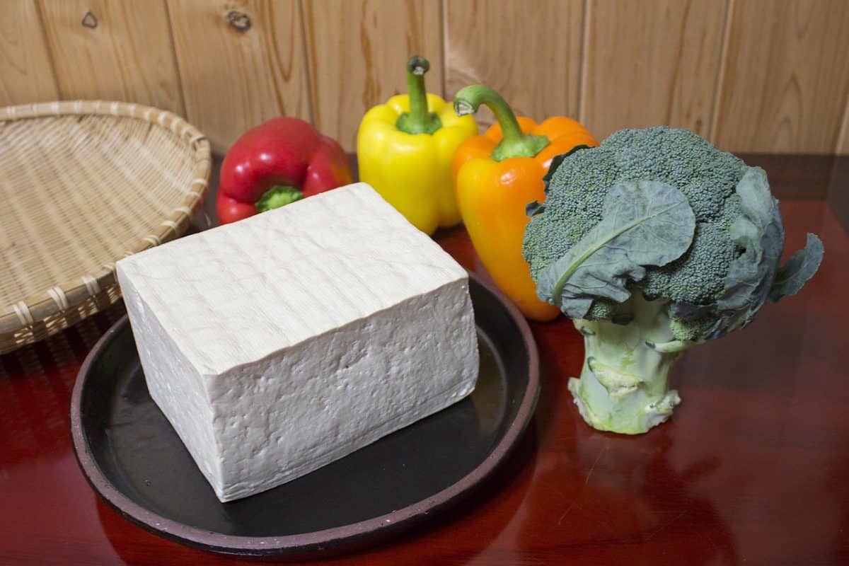 A block of tofu next to colorful bell peppers and broccoli.