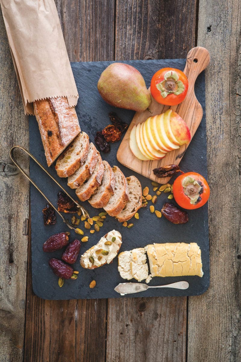 A rustic cheese board with tofu chevre, sliced bread, fruits, and nuts.