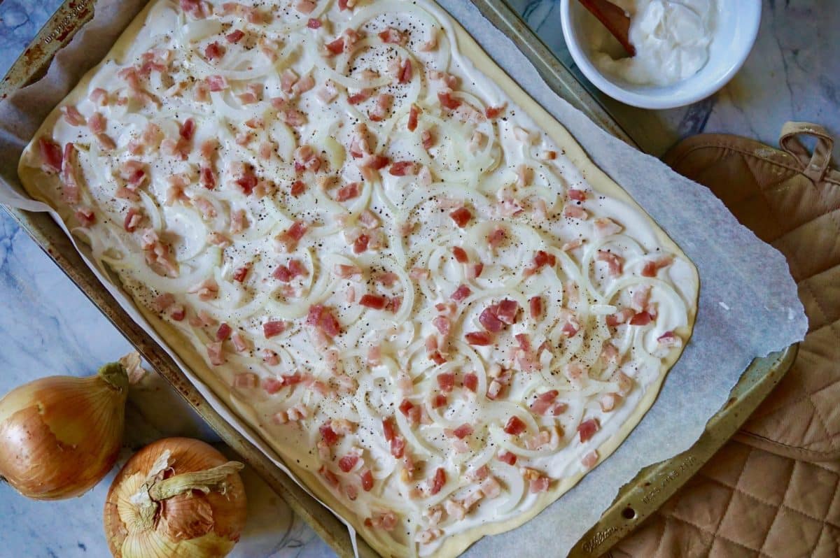 Uncooked flammkuchen with onions and sauce on a baking sheet.