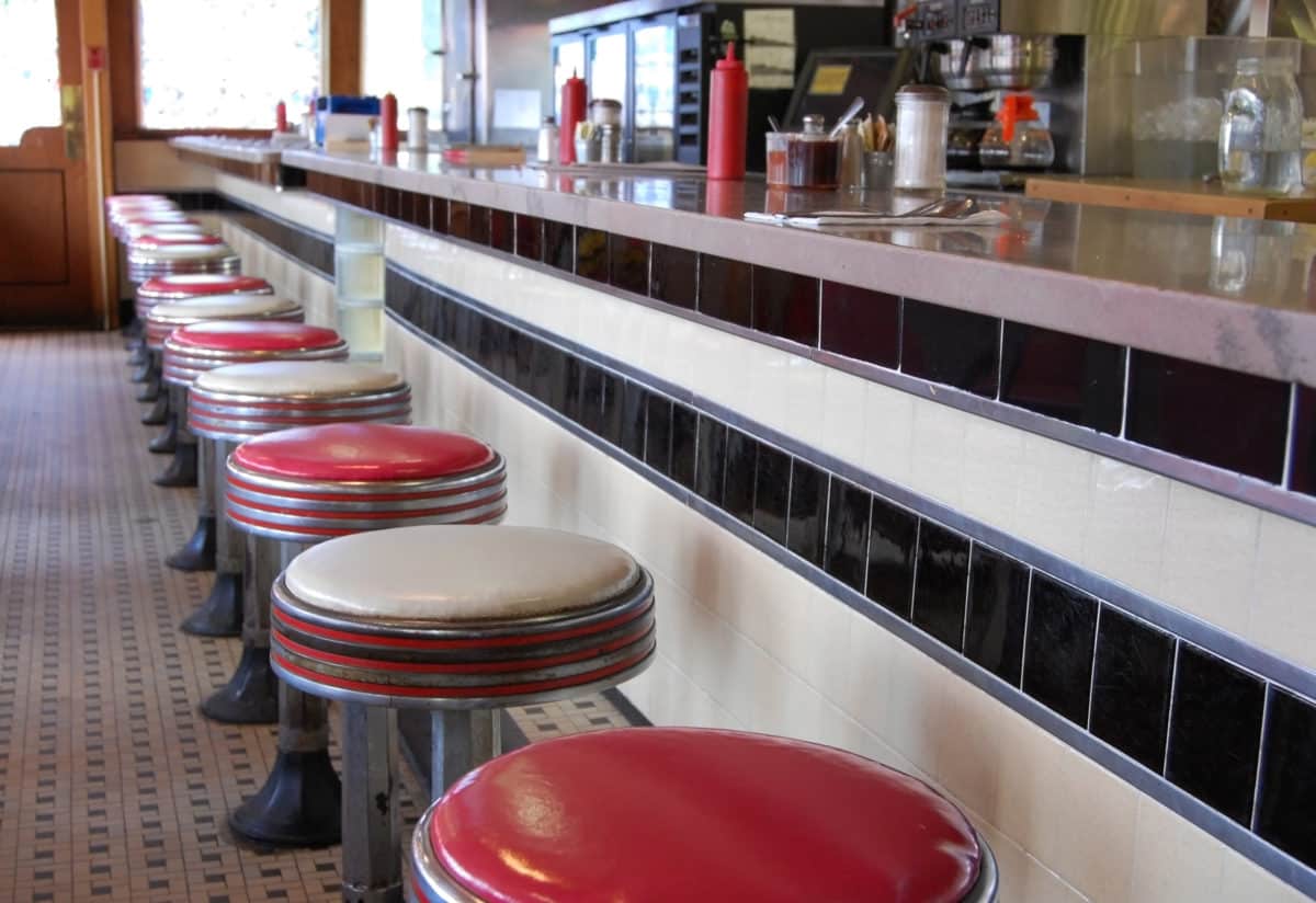 Interior of an old-fashioned diner with red art deco style bar stools, white tiled floor, and counter with condiments.