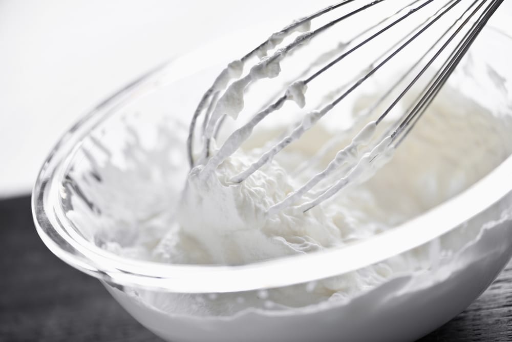 Close-up of a metal whisk whipping white cream in a clear glass bowl.